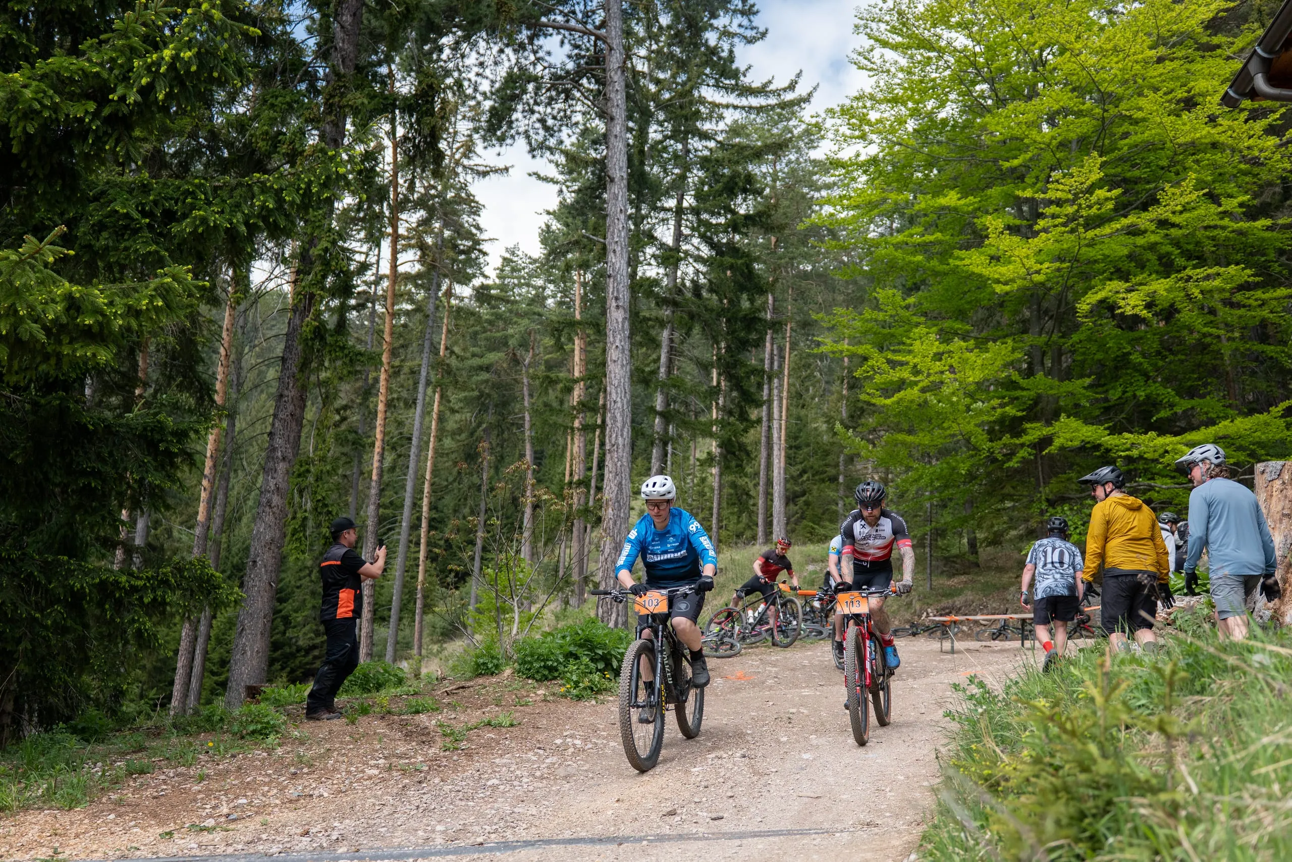 Fahrer kommen den breiten Forstweg im Wald herunter, am Rand ein Streckenposten mit oranger Weste