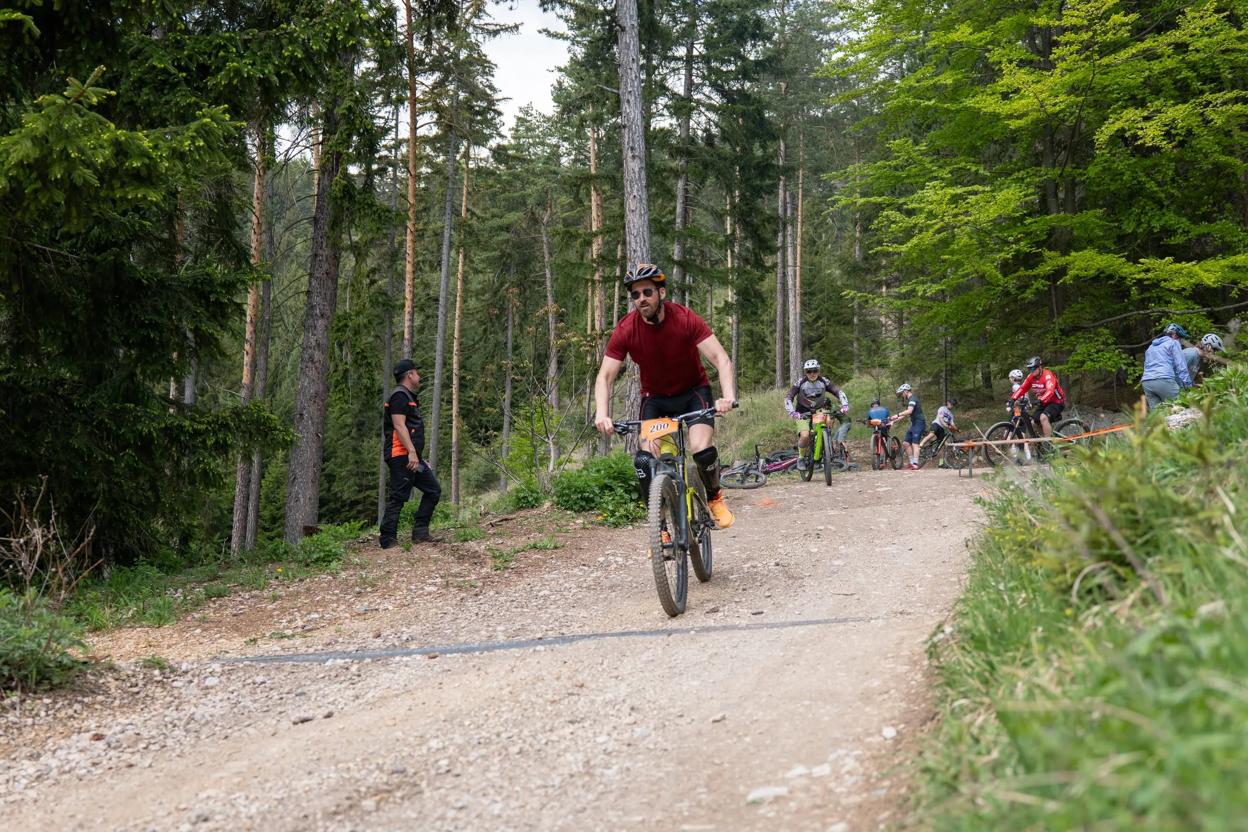 Fahrer kommen den breiten Forstweg im Wald herunter, am Rand ein Streckenposten mit oranger Weste