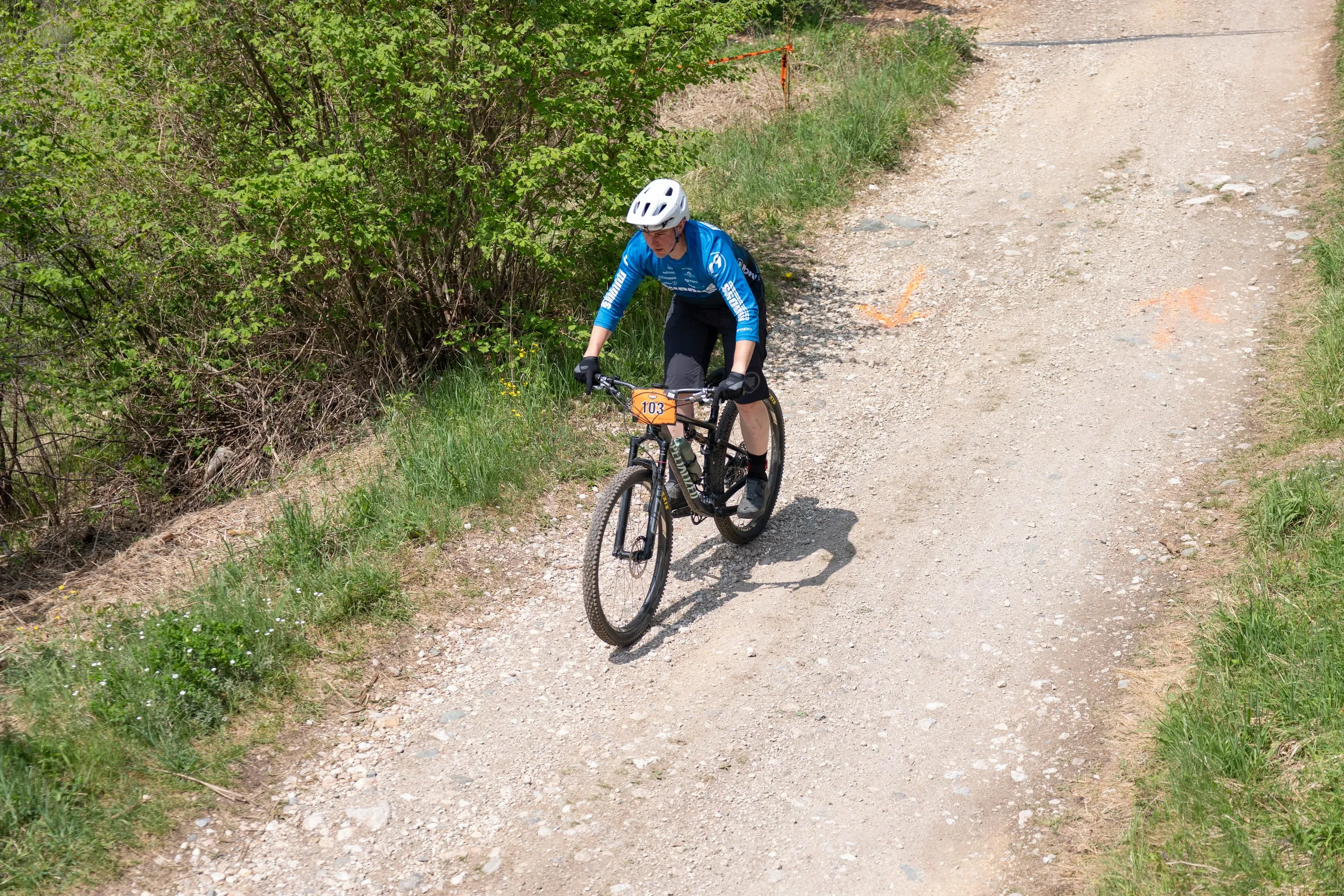 Ein Fahrer im blauen Trikot fährt eine Schotterkurve bergauf, die Sonne wirft harte Schatten